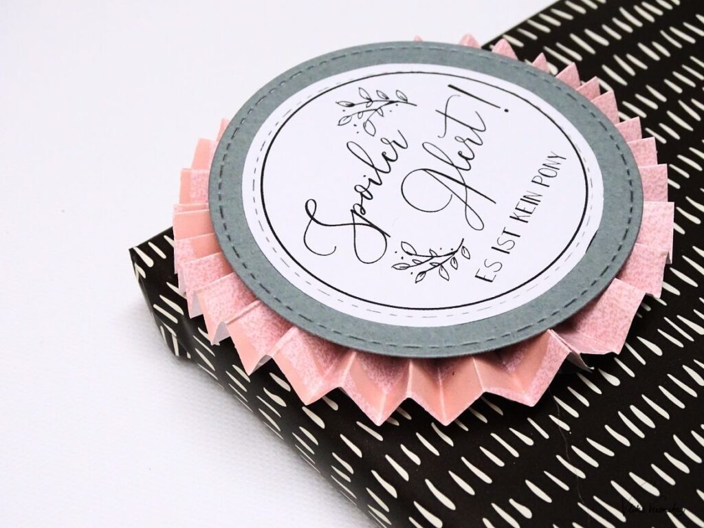 Bastle eine Rosette aus Papier und klebe einen runden Textbutton drauf.